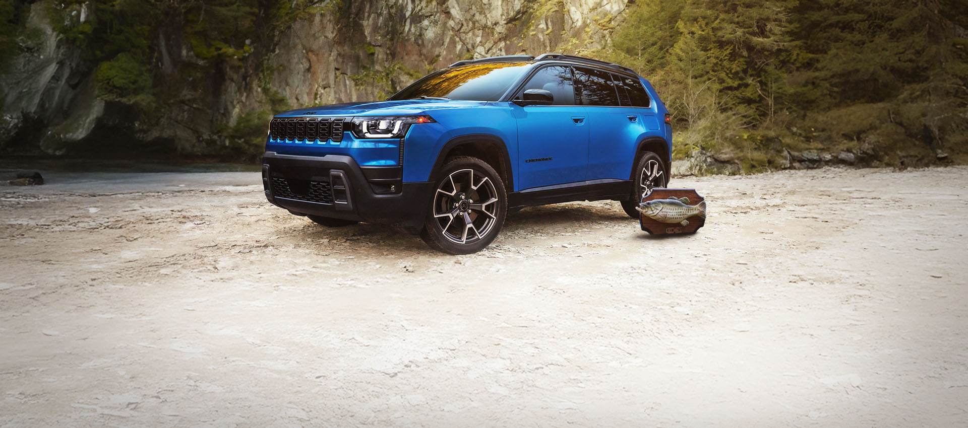 Vista del ángulo frontal del lado del conductor del Jeep Cherokee Overland 2026 azul estacionado en un claro, con un Billy Bass montado en la pared a su lado y un pantano boscoso de fondo.