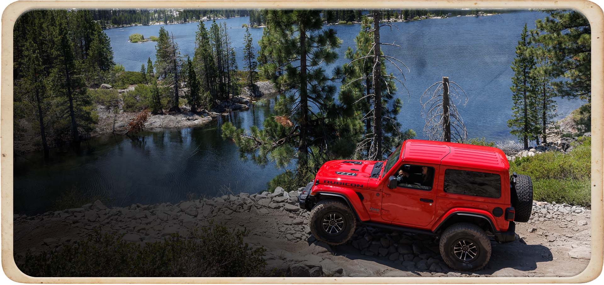 Un Jeep Wrangler Rubicon X 2025 rojo estacionado en un peñasco rocoso con vista a Lake Tahoe y a un bosque tupido.