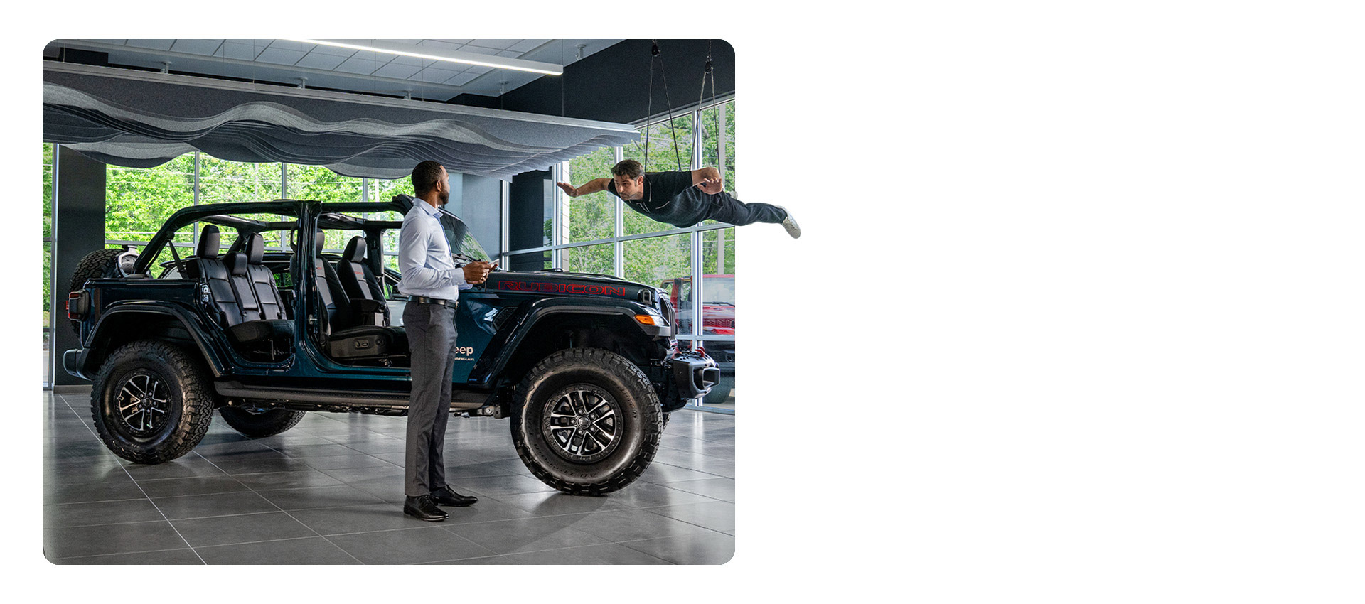Un Jeep Wrangler Rubicon X 2025 con sus puertas y techo extraídos en exhibición en la sala de un concesionario, con un hombre parado cerca y otro colgado de cables, flotando arriba.
