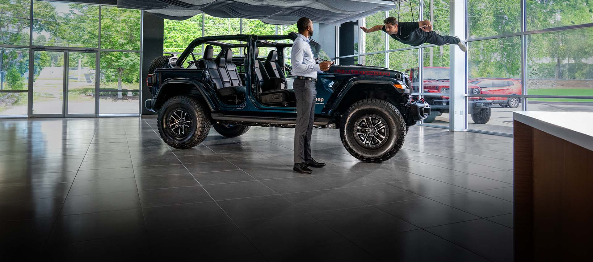 Un Jeep Wrangler Rubicon X 2025 con sus puertas y techo extraídos en exhibición en la sala de un concesionario, con un hombre parado cerca y otro colgado de cables, flotando arriba.