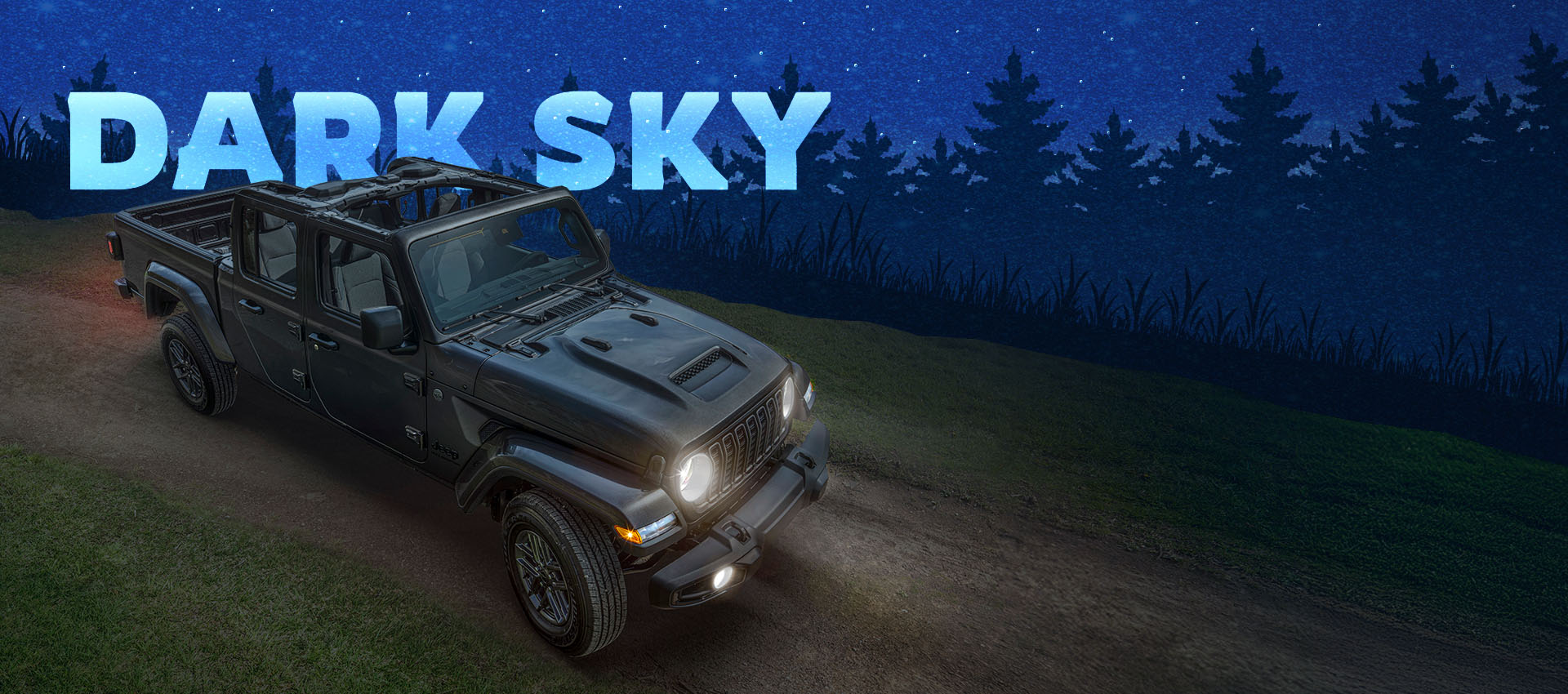 Ángulo delantero elevado del lado del pasajero de un Jeep Gladiator Sport S 2025 en gris oscuro con el paquete Dark Sky en un camino de tierra, con los faros delanteros encendidos y el techo extraído. Paquete Dark Sky.