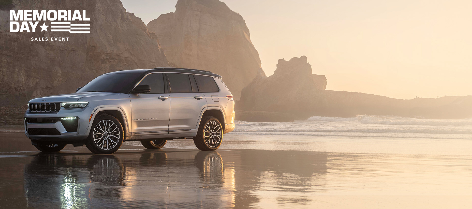 Un Jeep Grand Cherokee Summit 2026 plateado, estacionado en una playa, con formaciones rocosas al fondo.​​​​​​​ The Memorial Day Sales Event.