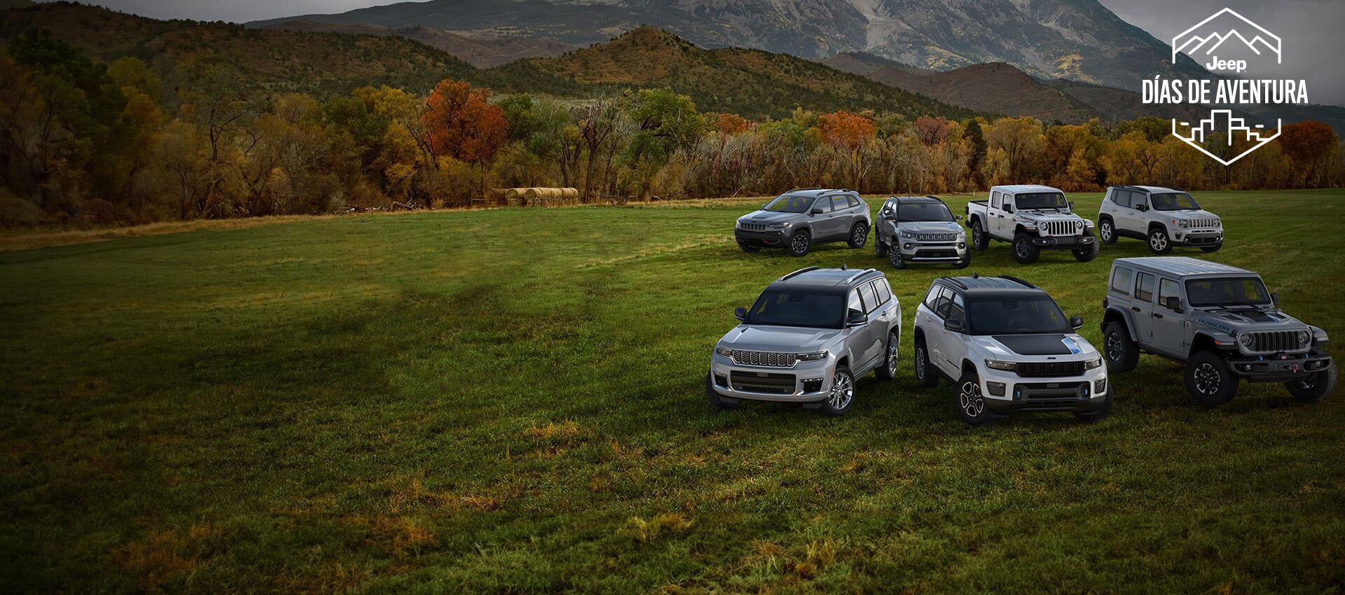 The Jeep Brand lineup parked in two rows on a grassy plain. Beyond is a forest wrapped in autumn leaves and mountains rising in the distance.