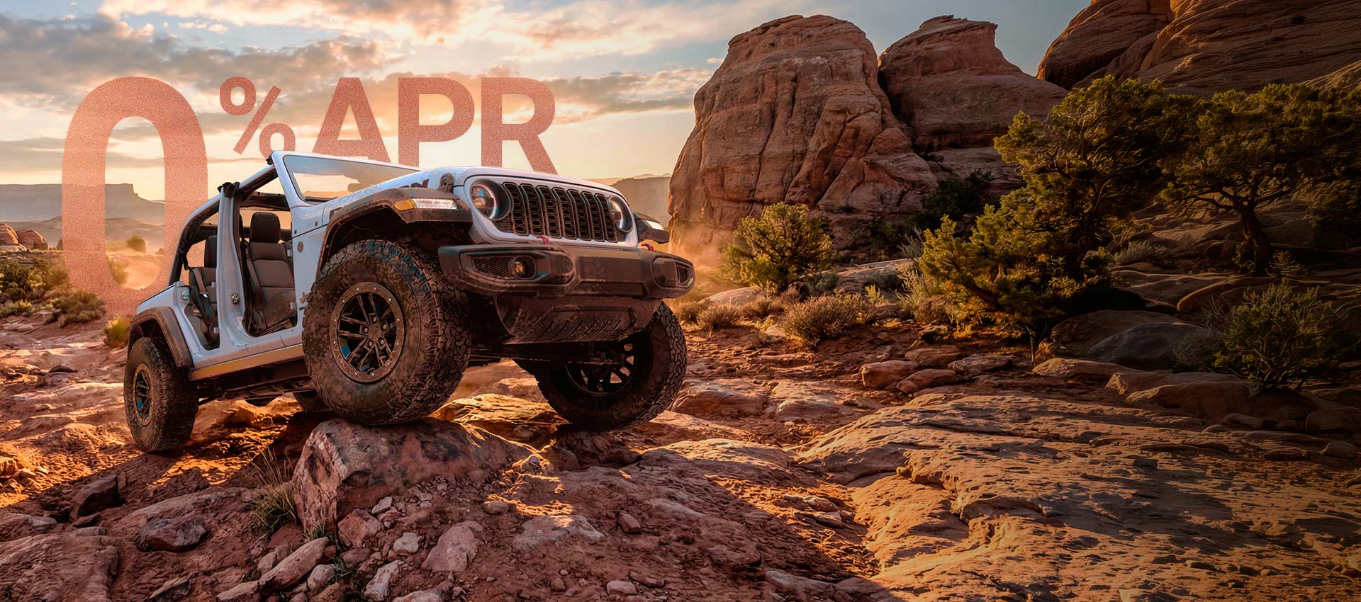 A white 2026 Jeep Wrangler Rubicon with its doors and top removed, crawling over a very rugged trail of bedrock. 0% APR. Drive Into Fall Sales Event.